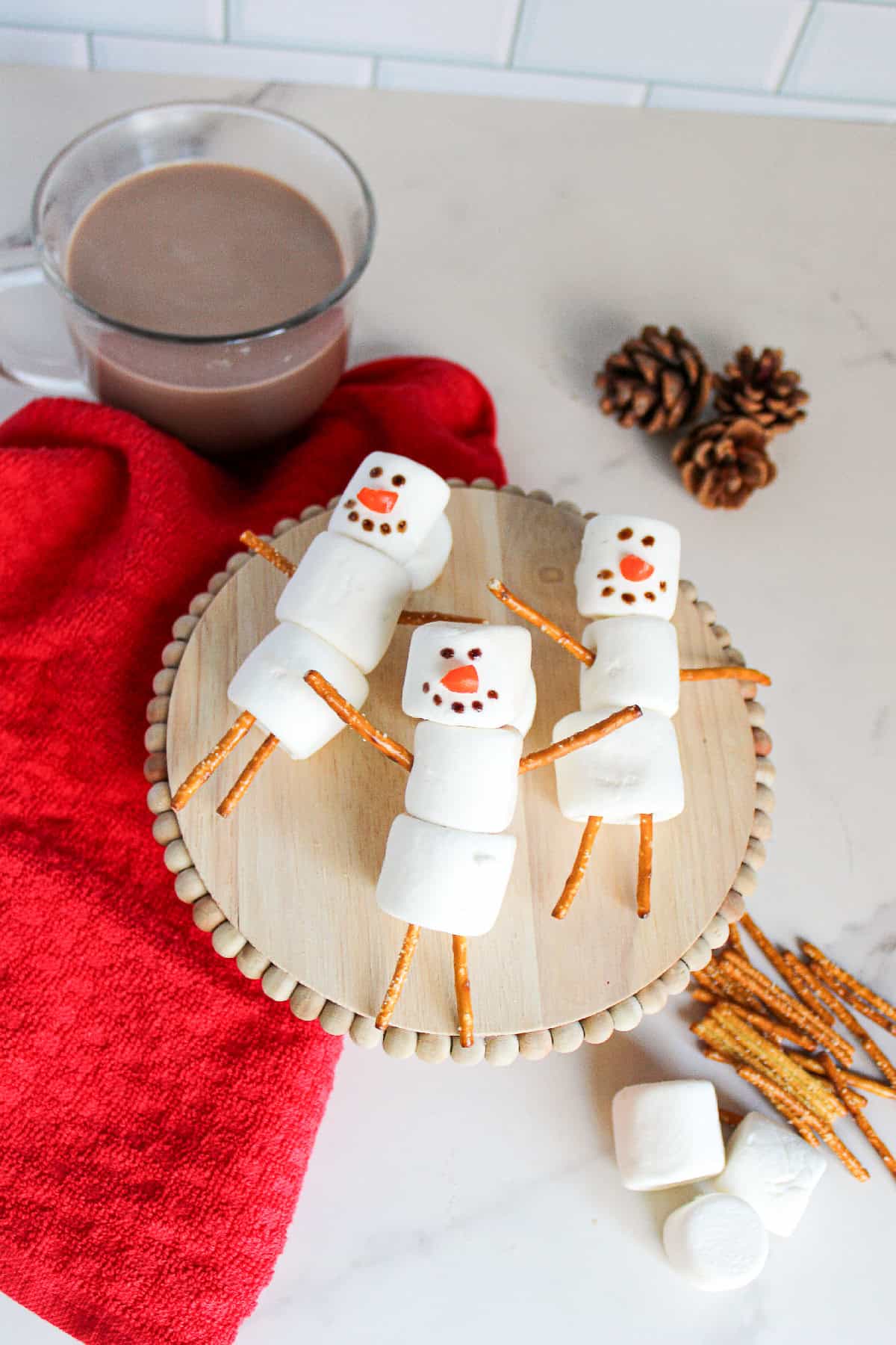 Far view of marshmallow snowmen with pretzel stick limbs and decorated faces on a wooden tray next to a mug of hot chocolate.