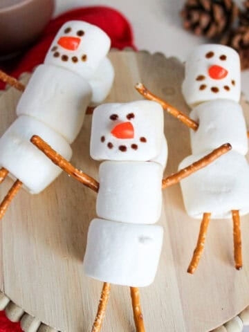 Three marshmallow snowmen with pretzel stick arms and legs arranged on a wooden stand, surrounded by a red towel, hot cocoa, and pinecones.