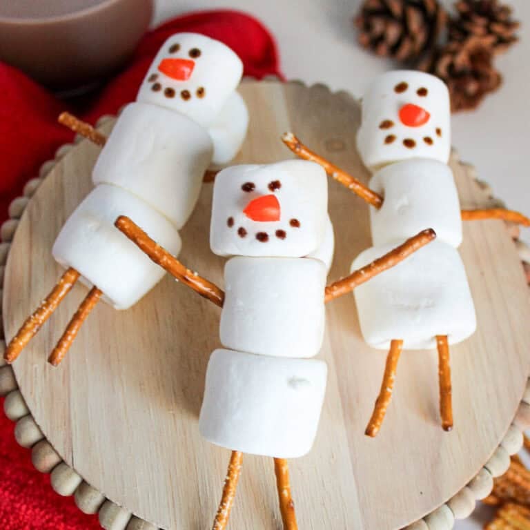 Three marshmallow snowmen with pretzel stick arms and legs arranged on a wooden stand, surrounded by a red towel, hot cocoa, and pinecones.
