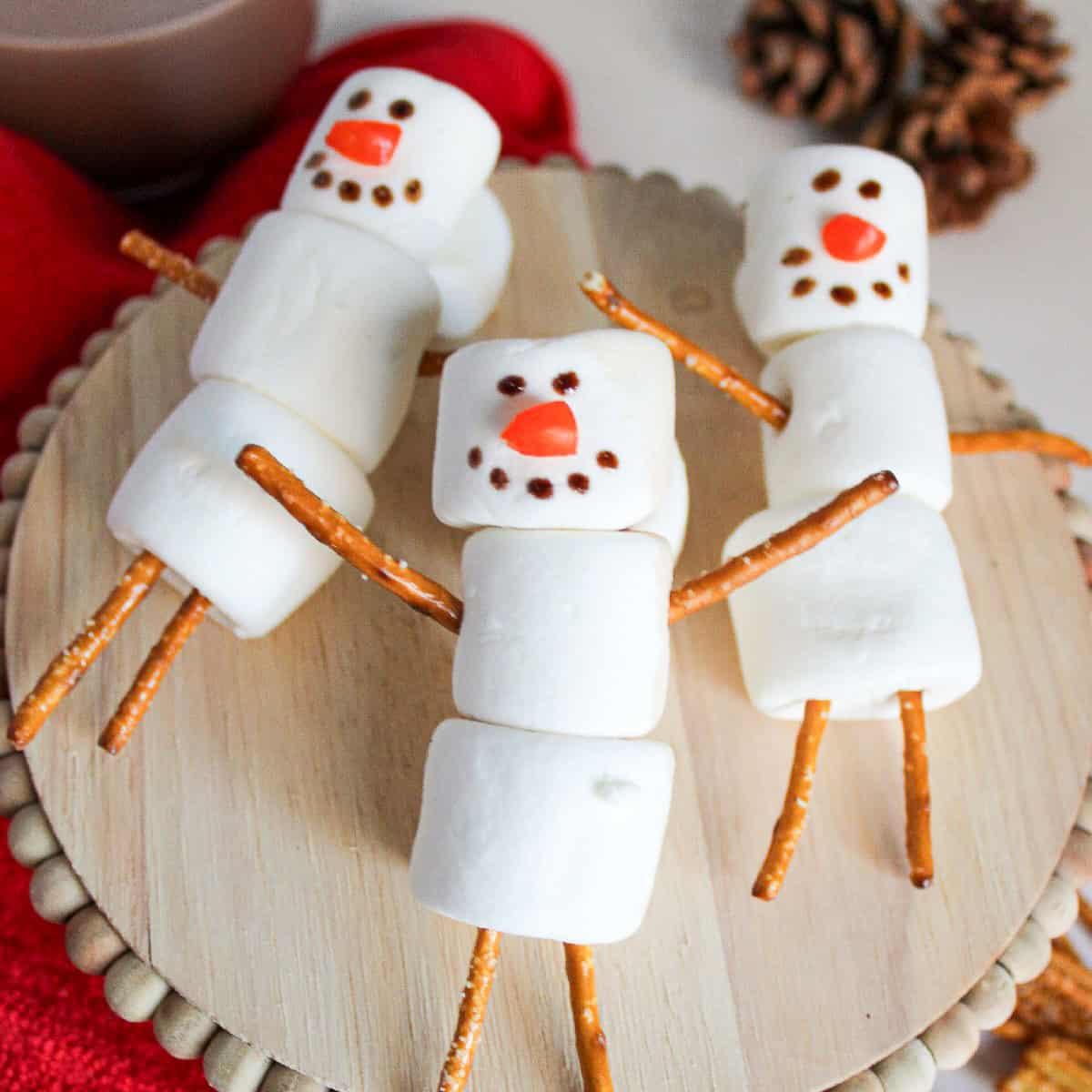 Three marshmallow snowmen with pretzel stick arms and legs arranged on a wooden stand, surrounded by a red towel, hot cocoa, and pinecones.