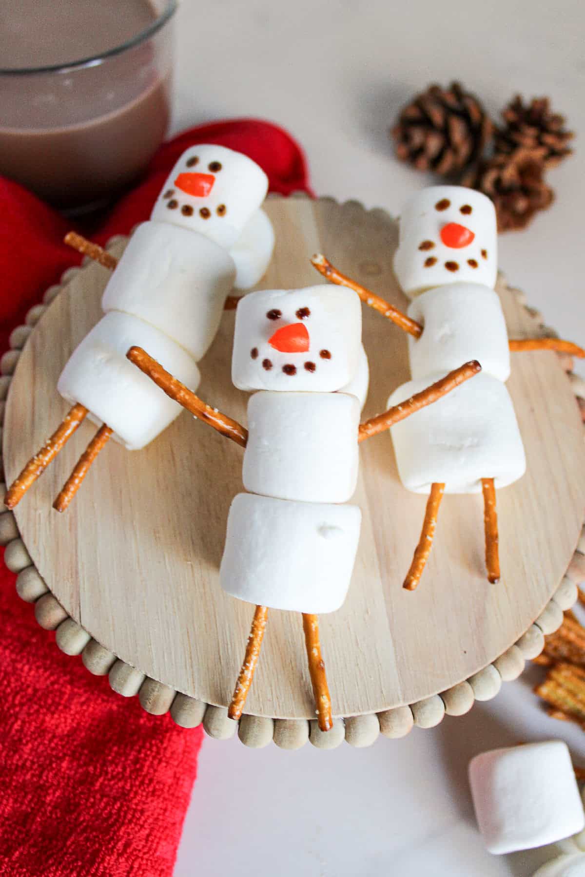 Top view of marshmallow snowmen with pretzel arms and legs, festive red towel, and hot chocolate in the background.