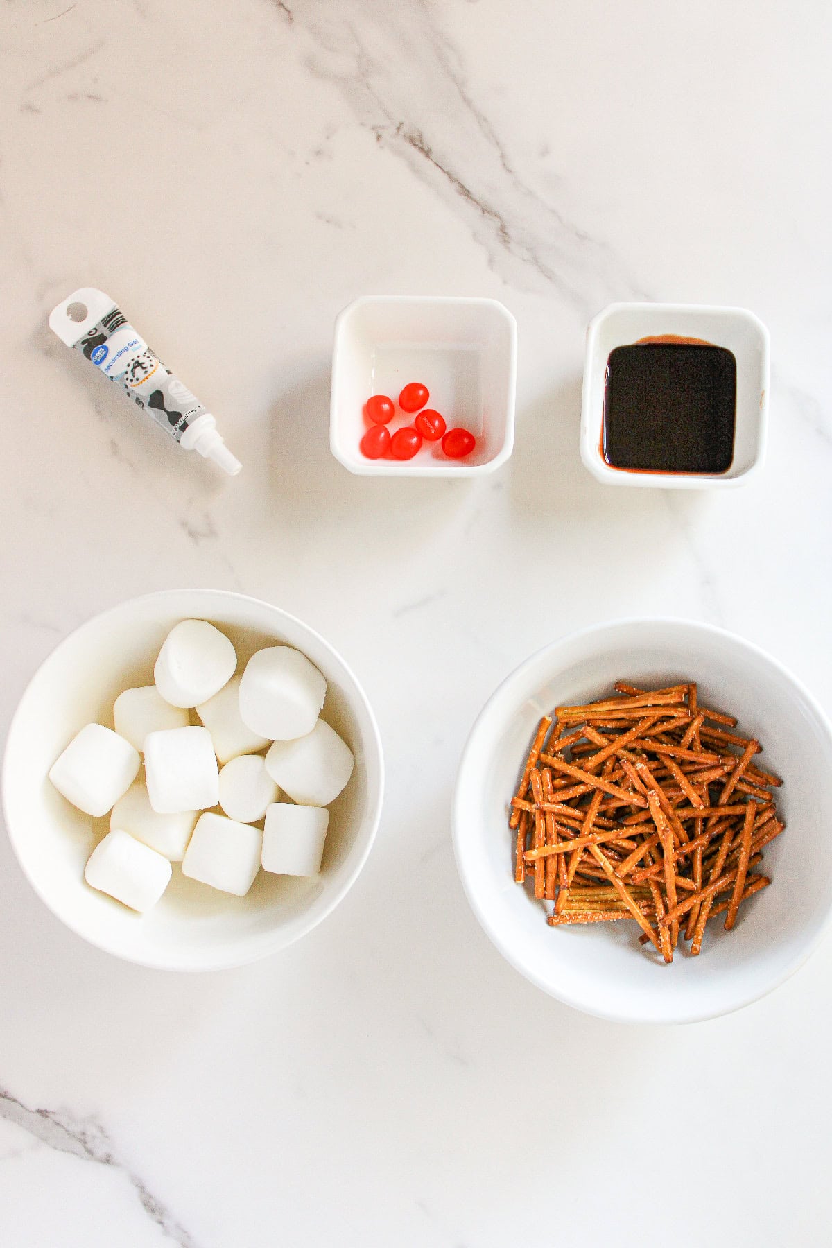 Marshmallows, pretzel sticks, red jelly beans, chocolate syrup, and black decorating gel arranged in bowls on a white marble surface.