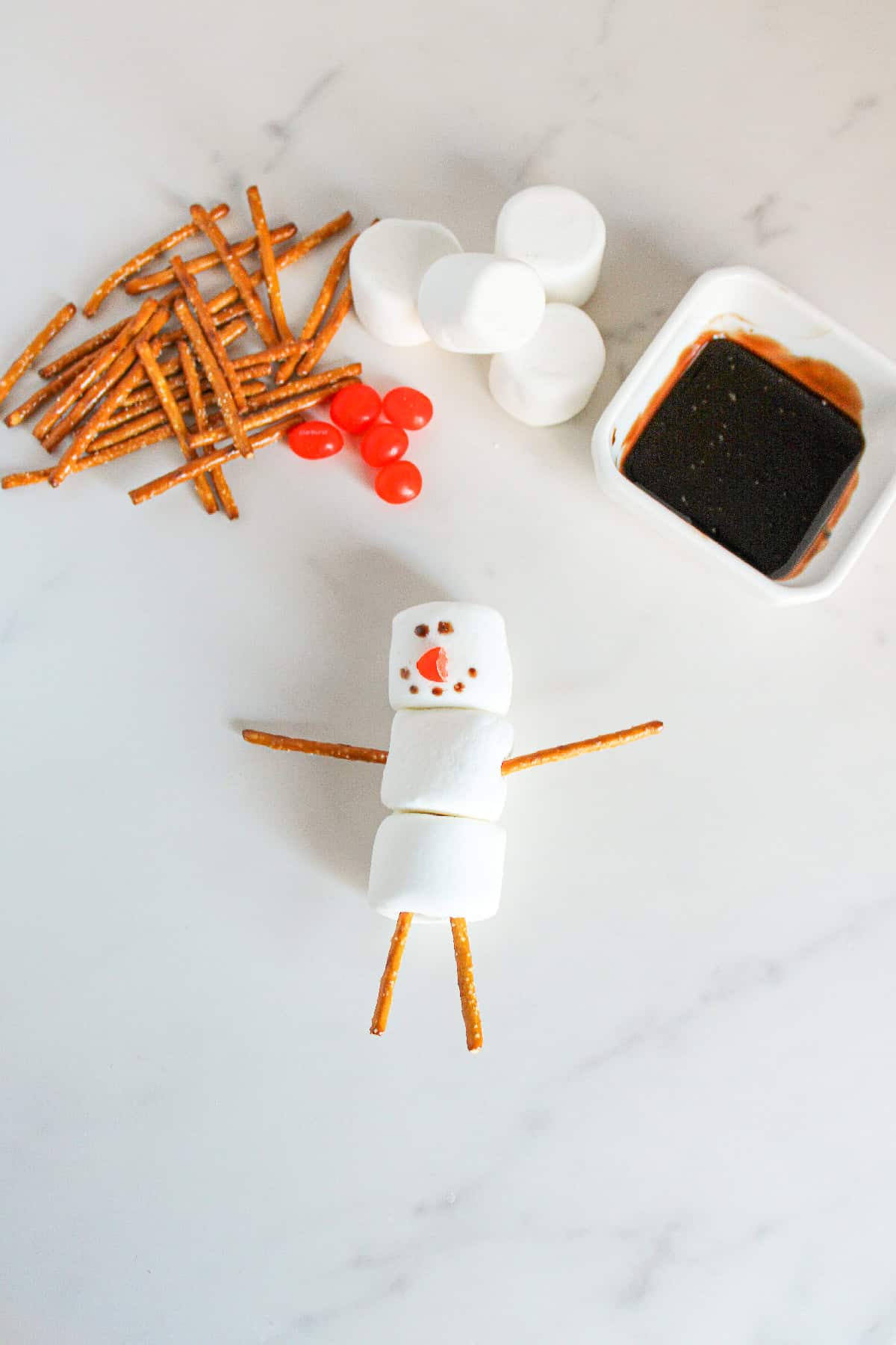 Completed marshmallow snowman with chocolate eyes and buttons, red nose, and pretzel arms and legs.