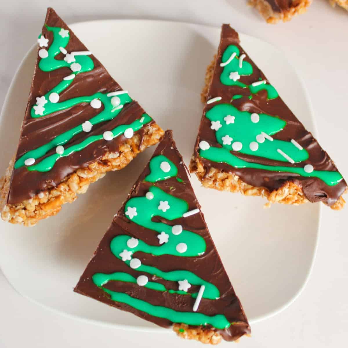 Close-up of three Christmas tree–shaped peanut butter bars on a white plate, topped with melted chocolate, green icing drizzle, and white sprinkles, creating a festive holiday dessert.