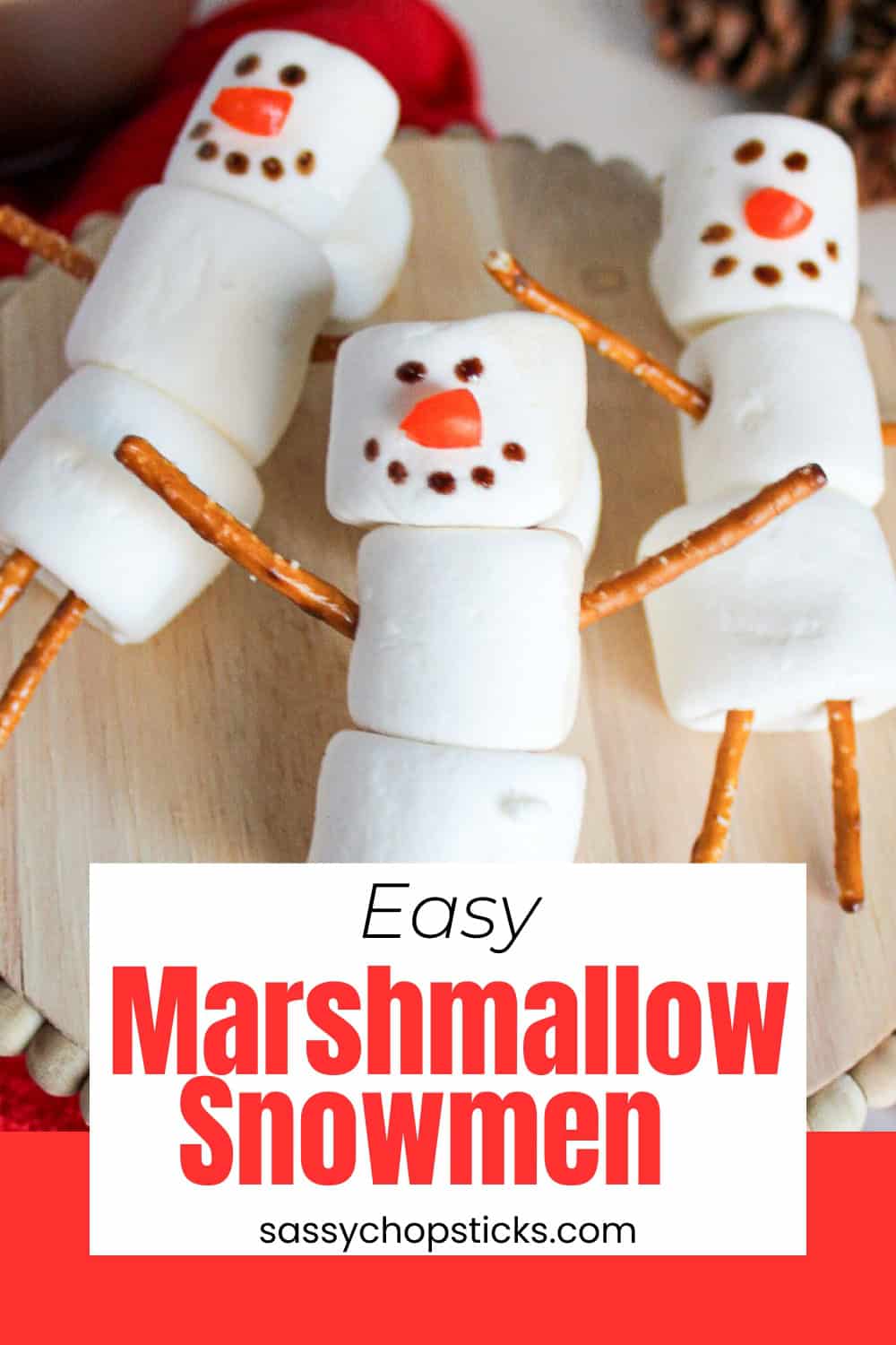 Three marshmallow snowmen with pretzel stick arms and legs arranged on a wooden stand, surrounded by a red towel, hot cocoa.