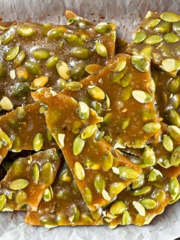 Pumpkin seed brittle pieces stacked on parchment paper, showing glossy caramel coating and green pumpkin seeds.