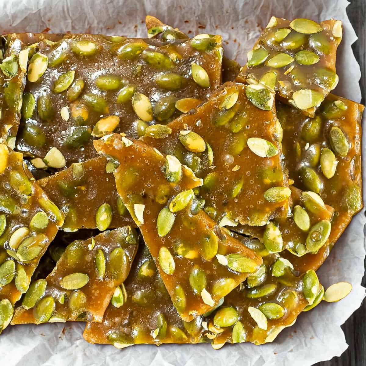 Pumpkin seed brittle pieces stacked on parchment paper, showing glossy caramel coating and green pumpkin seeds.
