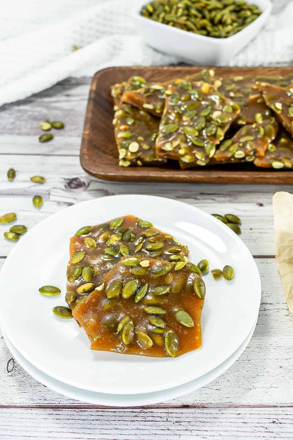 Pumpkin seed brittle pieces on a white plate with more brittle on a wooden tray in the background.