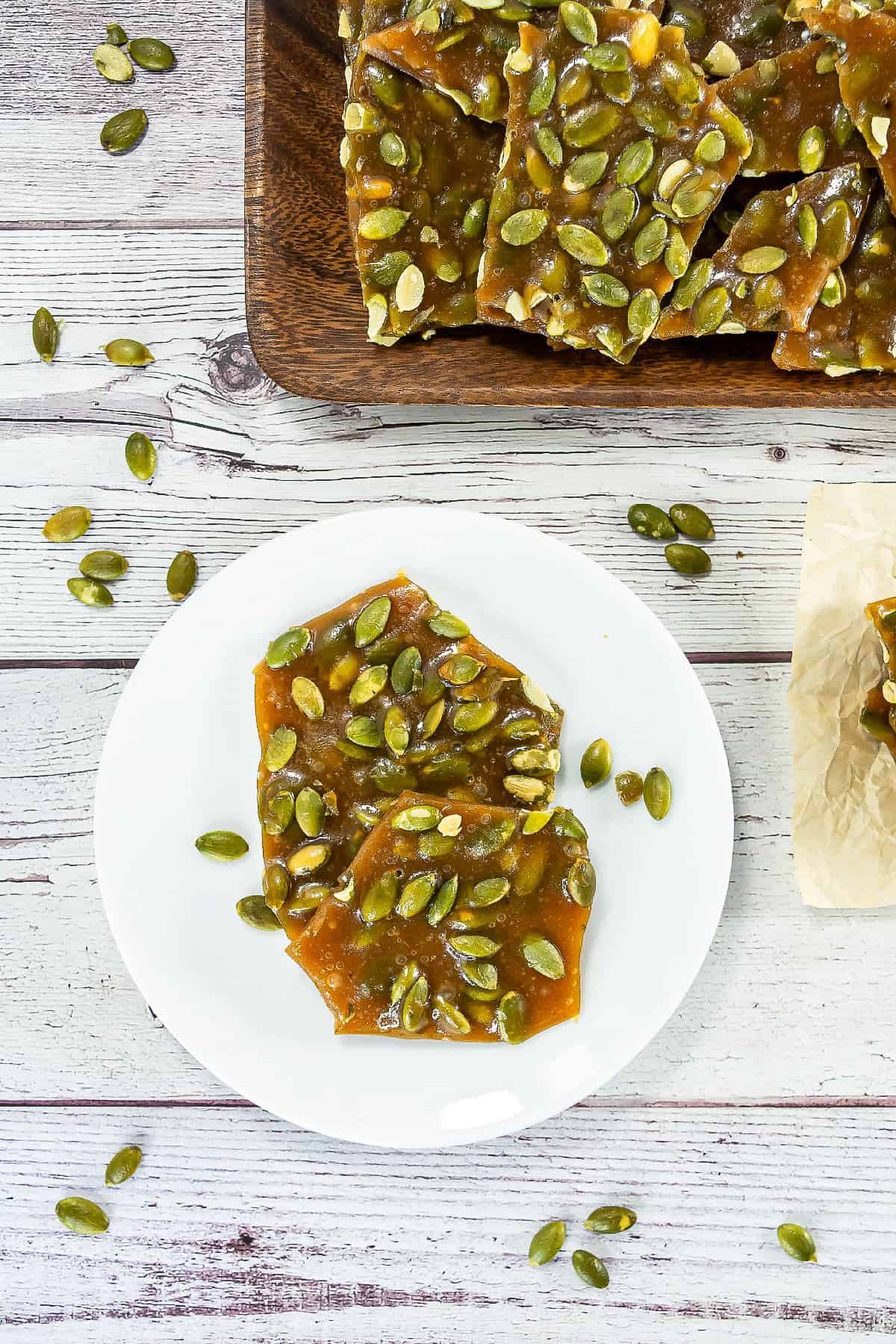 Top view of pumpkin seed brittle pieces on a white plate surrounded by scattered pepitas.