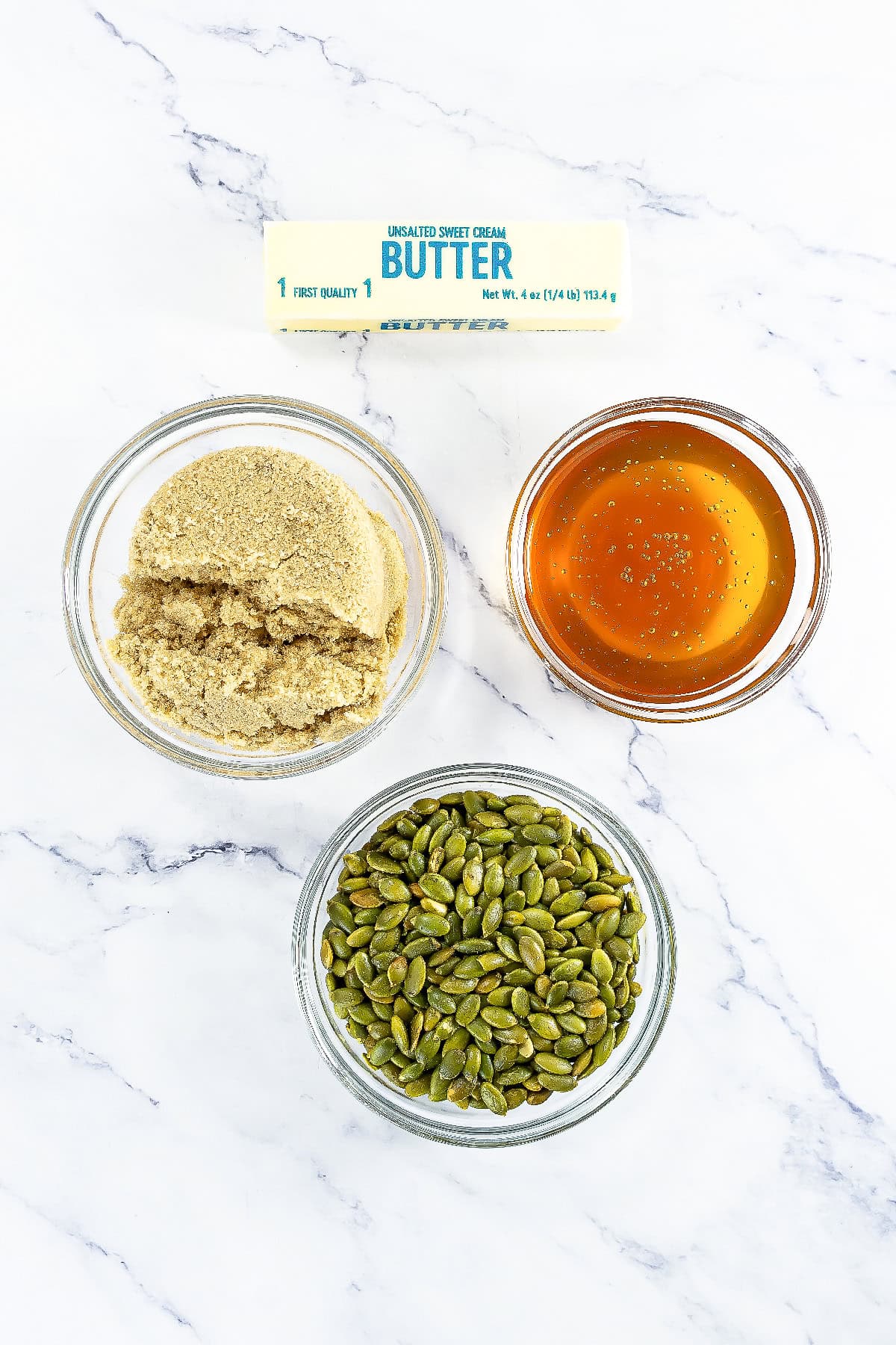 Top view of ingredients for pumpkin seed brittle including butter, brown sugar, honey, and pepitas arranged on a marble surface.