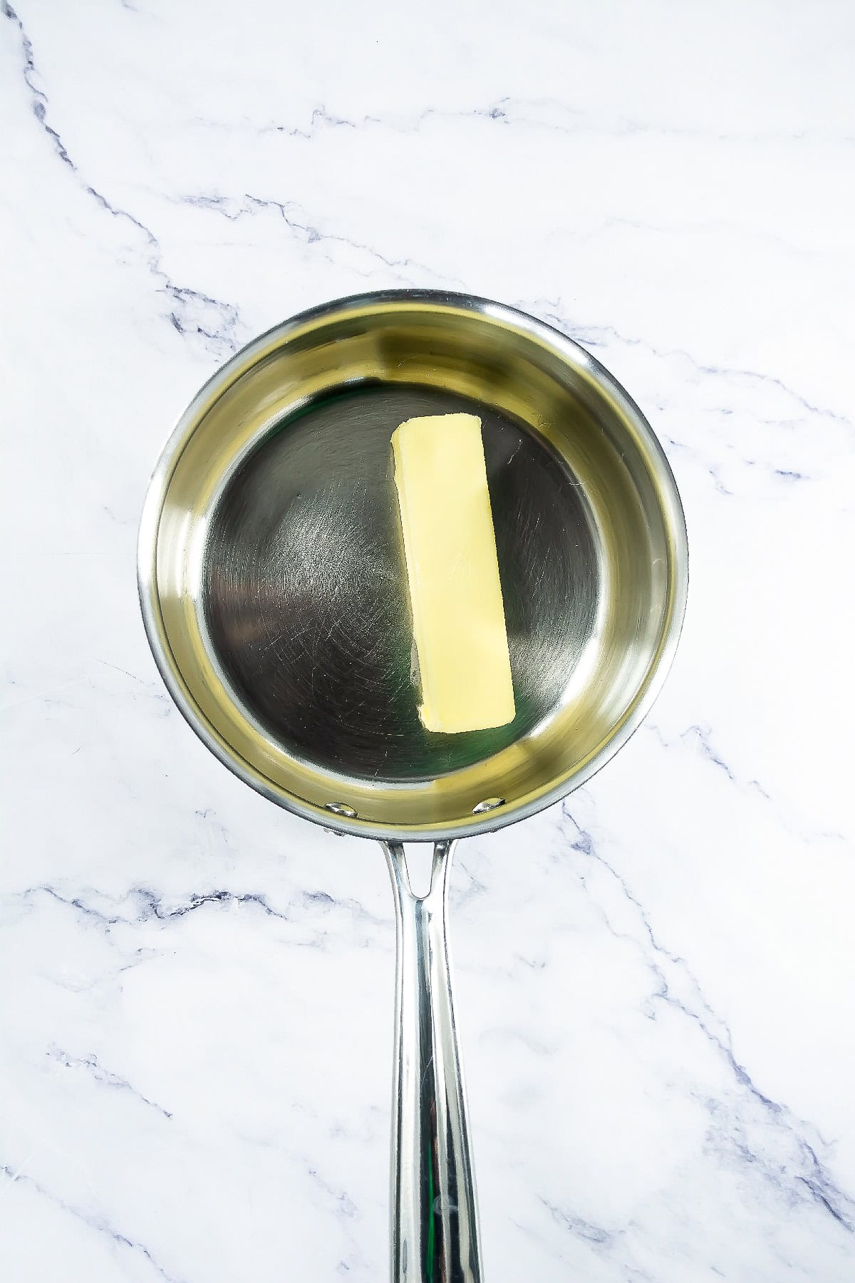 Stick of butter melting in a stainless steel saucepan on a marble countertop.