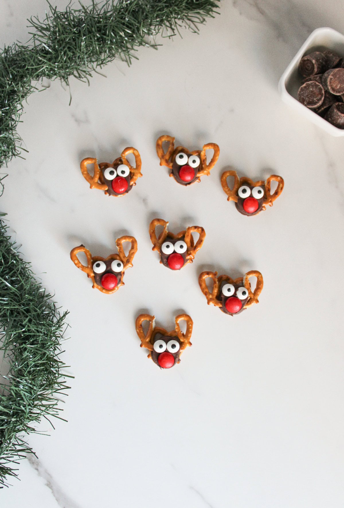Seven Rolo pretzel reindeer treats spread out on a white surface, surrounded by festive greenery and a small bowl of chocolates.