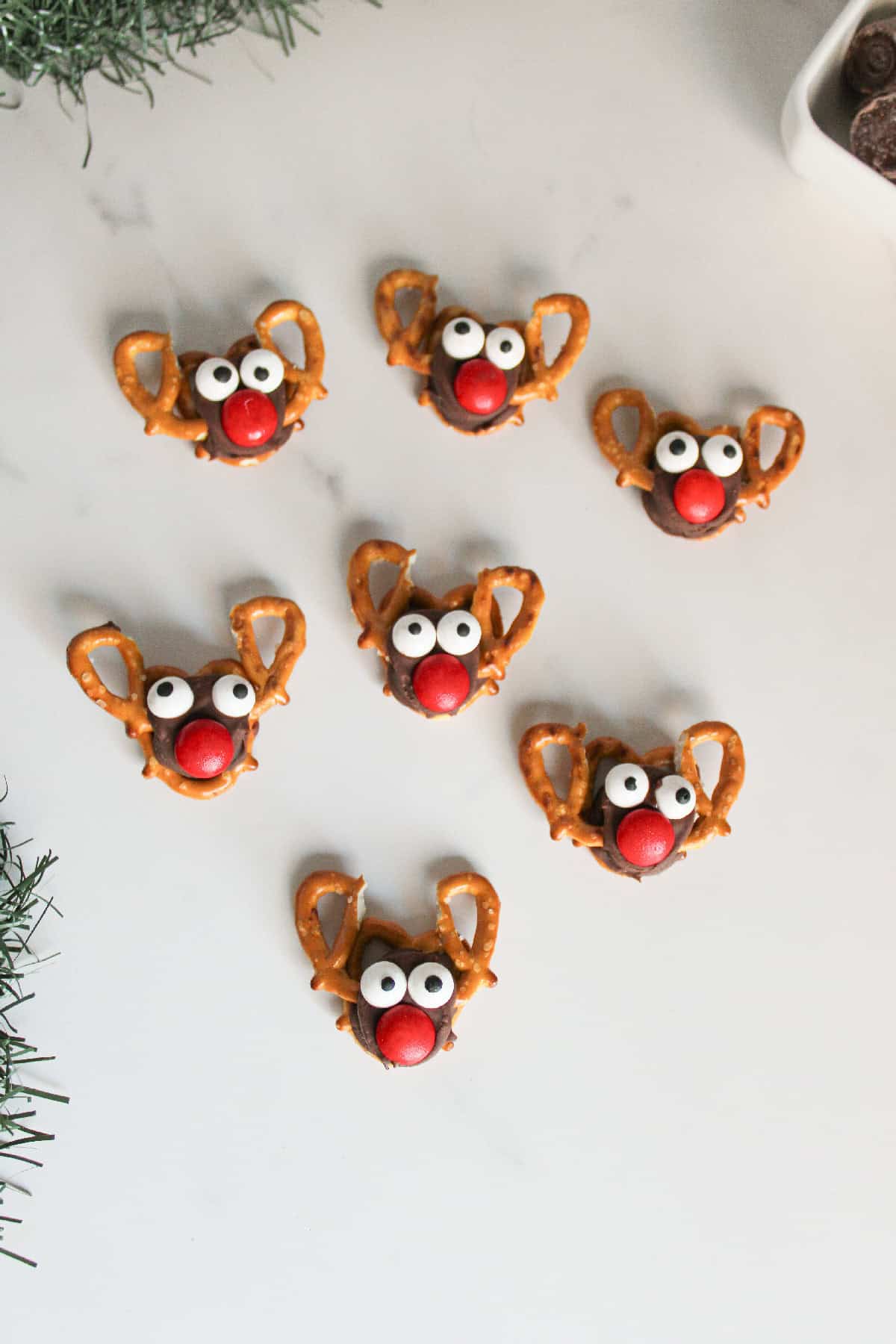 Close-up overhead view of Rolo pretzel reindeer treats with candy eyes and red noses, arranged playfully on a white marble background.