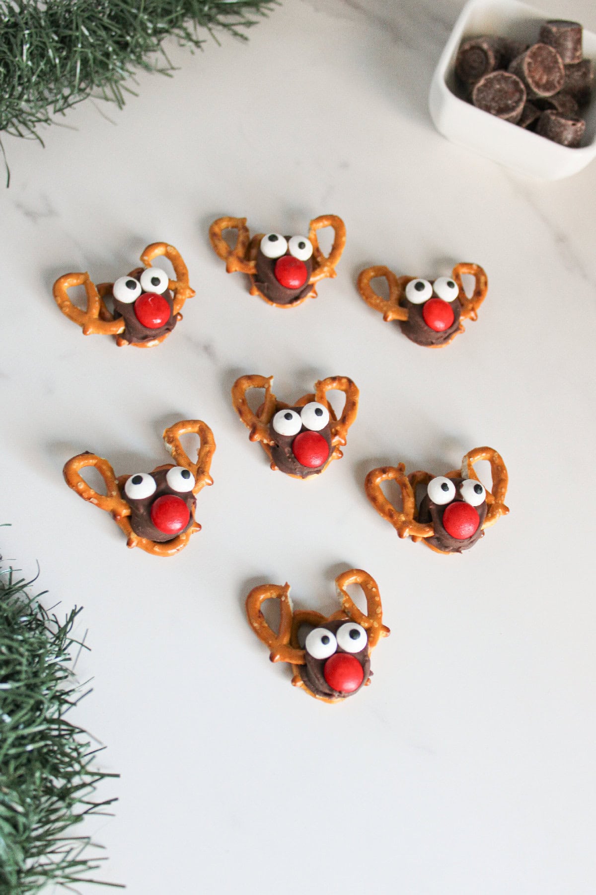 Rolo pretzel reindeer with candy eyes and red noses displayed in a scattered arrangement on a marble surface with holiday greenery.