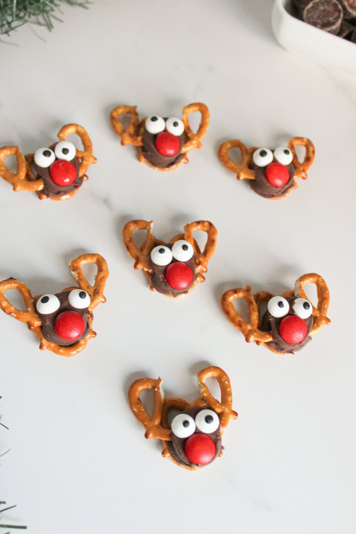 Rolo pretzel reindeer candies arranged in a light festive setting with greenery and chocolates in the background.