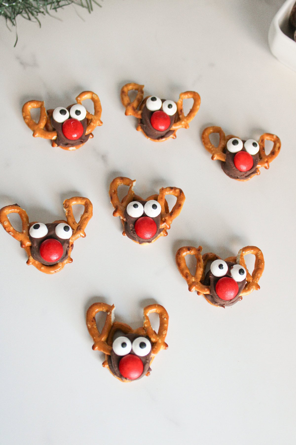 Overhead shot of several Rolo pretzel reindeer treats on a white background with green holiday garland framing the scene.