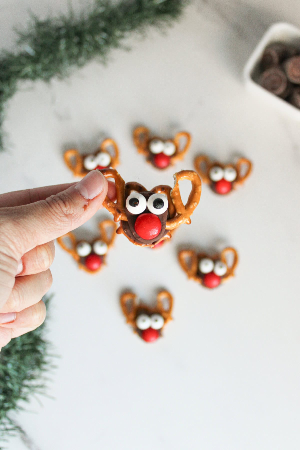 Close-up of a hand holding one Rolo pretzel reindeer treat with candy eyes and a red nose, with more treats blurred in the background.