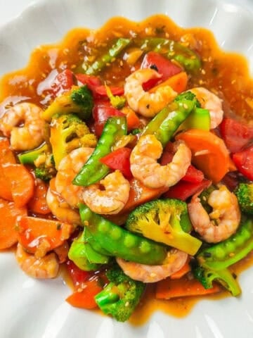 A colorful shrimp stir-fry served on a white scalloped plate, featuring plump shrimp, bright broccoli florets, snow peas, carrots, and red bell peppers coated in a glossy savory sauce.