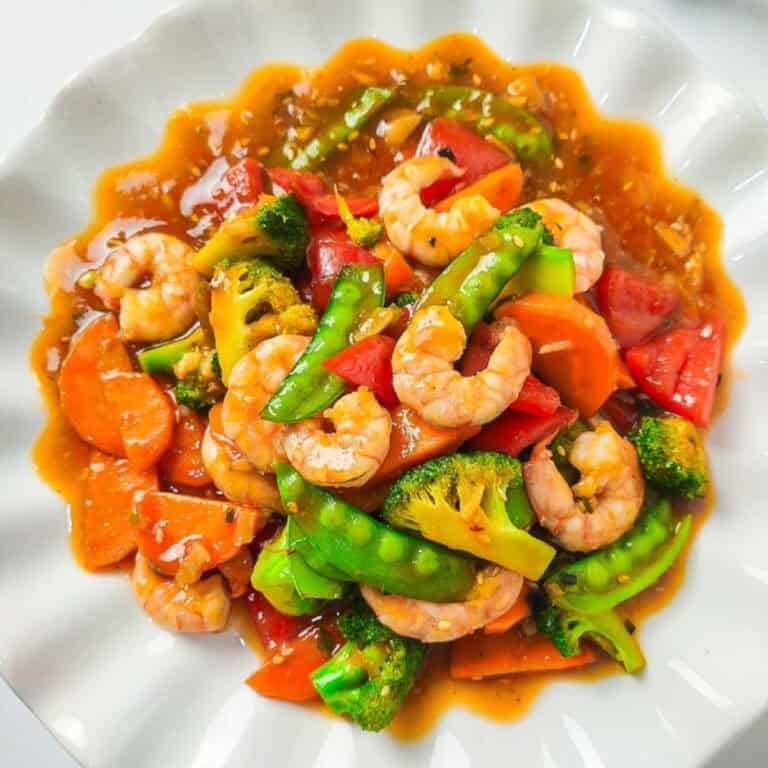 A colorful shrimp stir-fry served on a white scalloped plate, featuring plump shrimp, bright broccoli florets, snow peas, carrots, and red bell peppers coated in a glossy savory sauce.