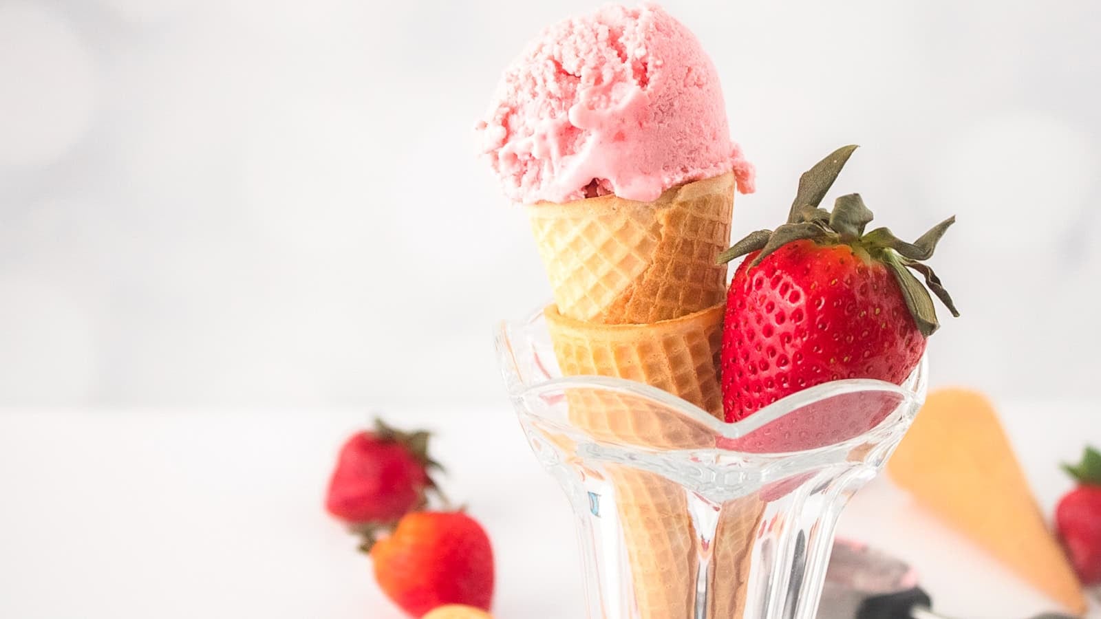 Vegan strawberry ice cream scooped into a waffle cone, served in a glass cup with fresh strawberries around it.