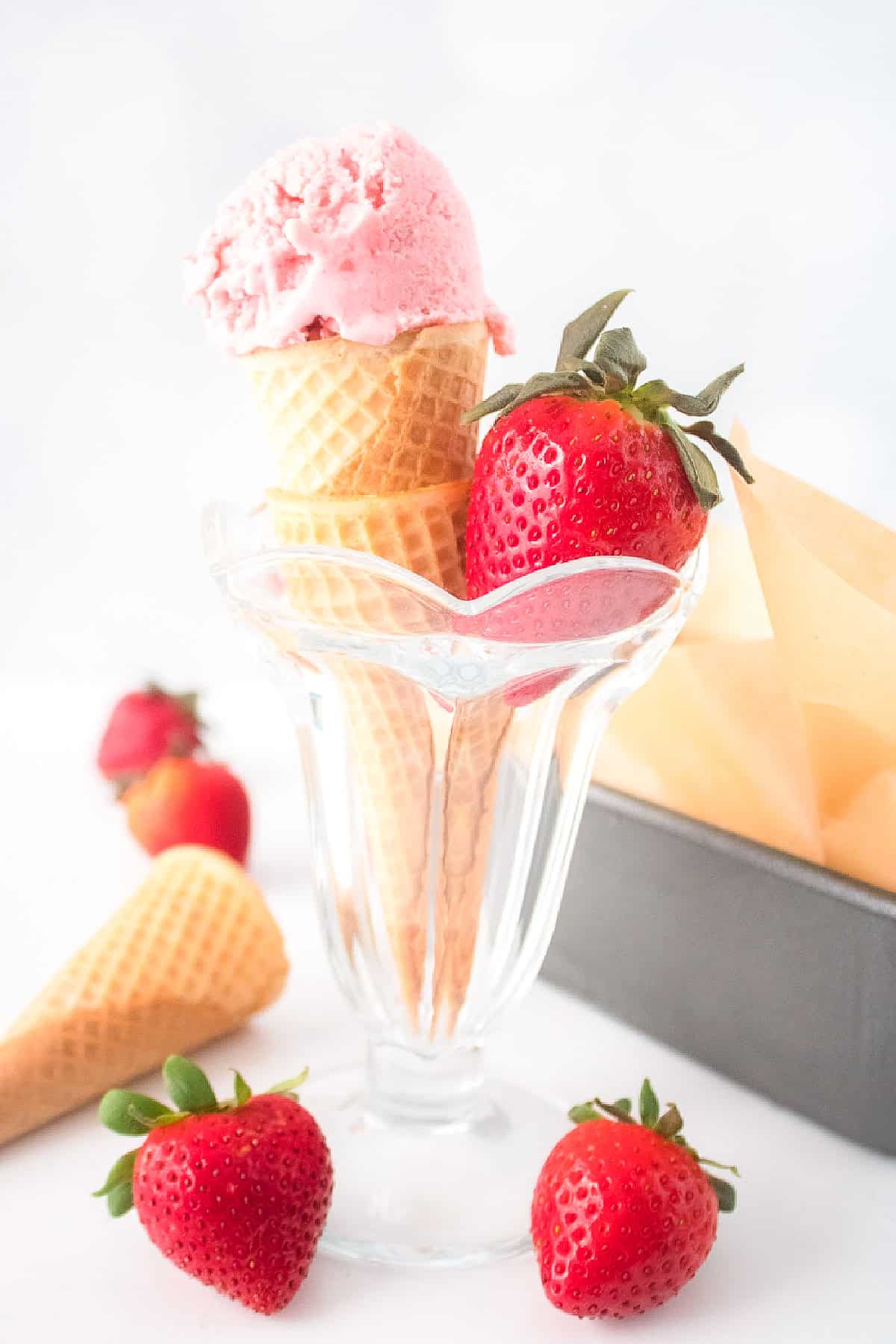 Vegan strawberry ice cream cone in a glass cup with fresh strawberries and waffle cones in the background.