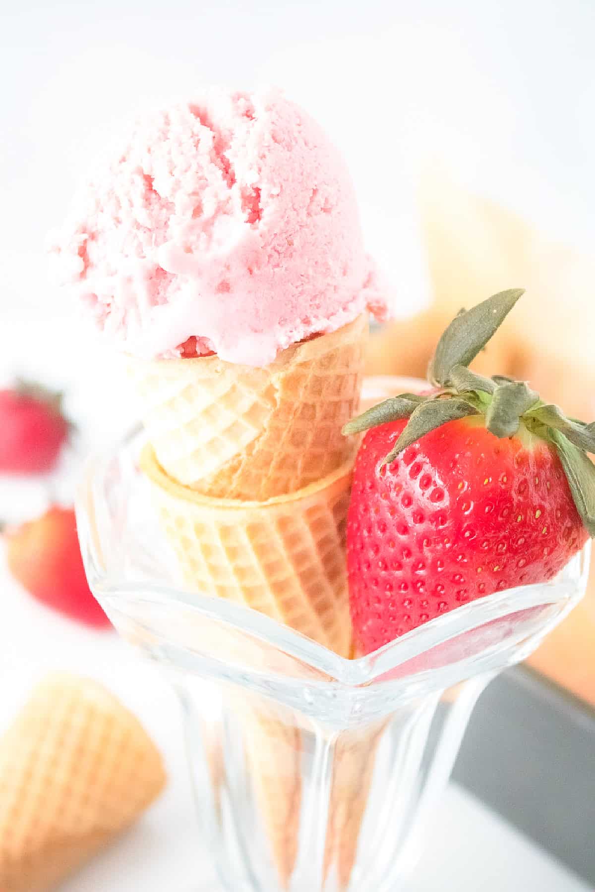 Vegan strawberry ice cream cone with fresh strawberry in a clear dessert glass, surrounded by waffle cones.
