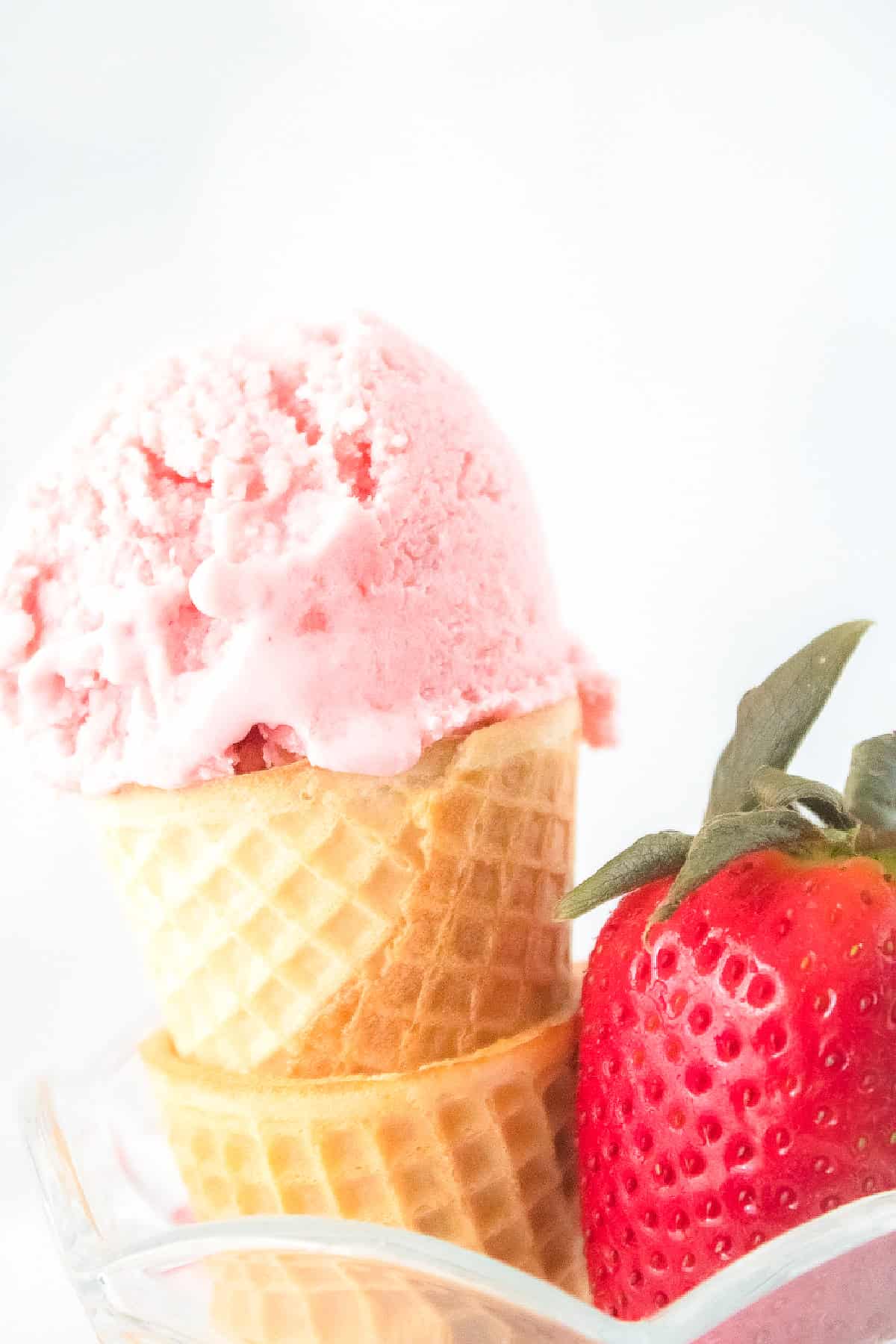 Close-up of a scoop of vegan strawberry ice cream in a waffle cone with a fresh strawberry beside it.