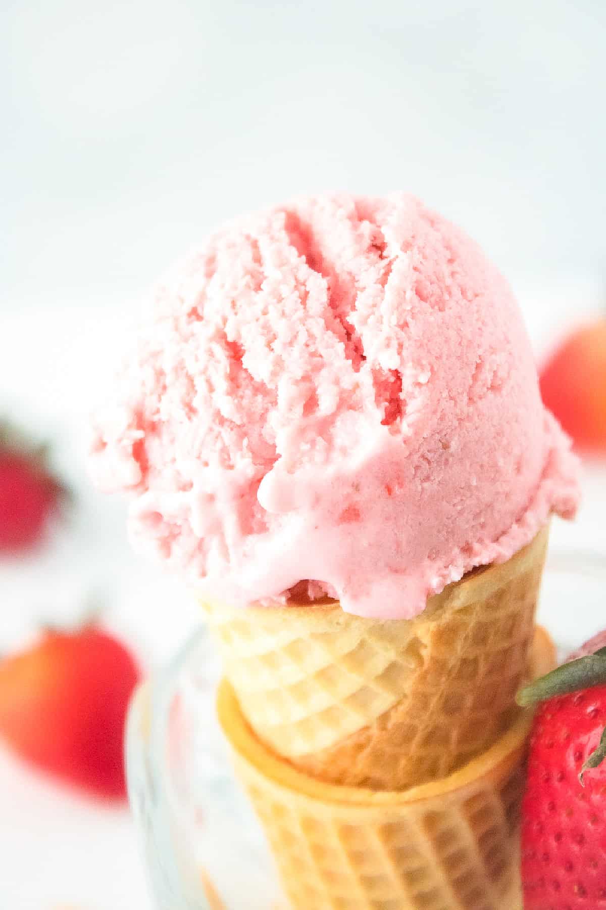 Vegan strawberry ice cream cone with fresh strawberries in the background on a bright white surface.