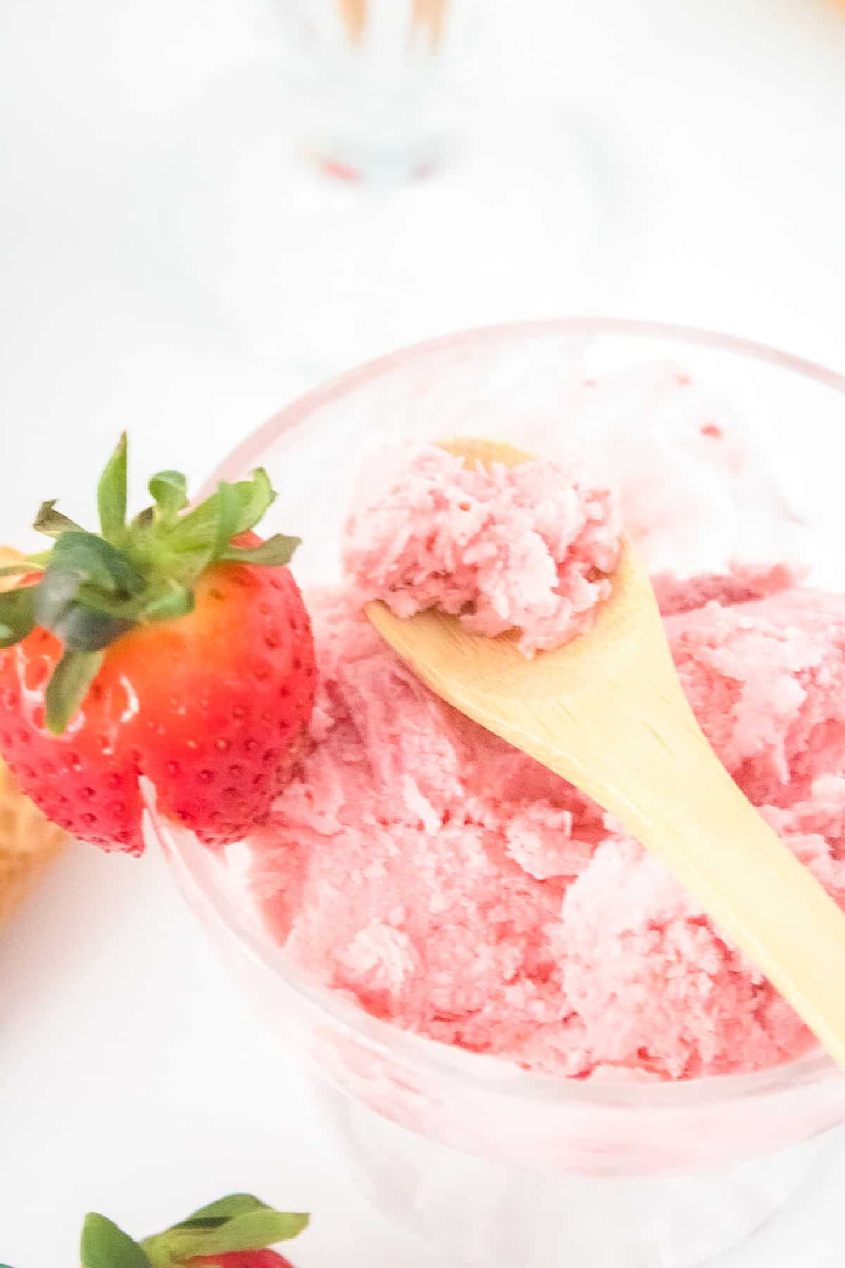 Bowl of creamy vegan strawberry ice cream with a wooden spoon and a fresh strawberry on top.