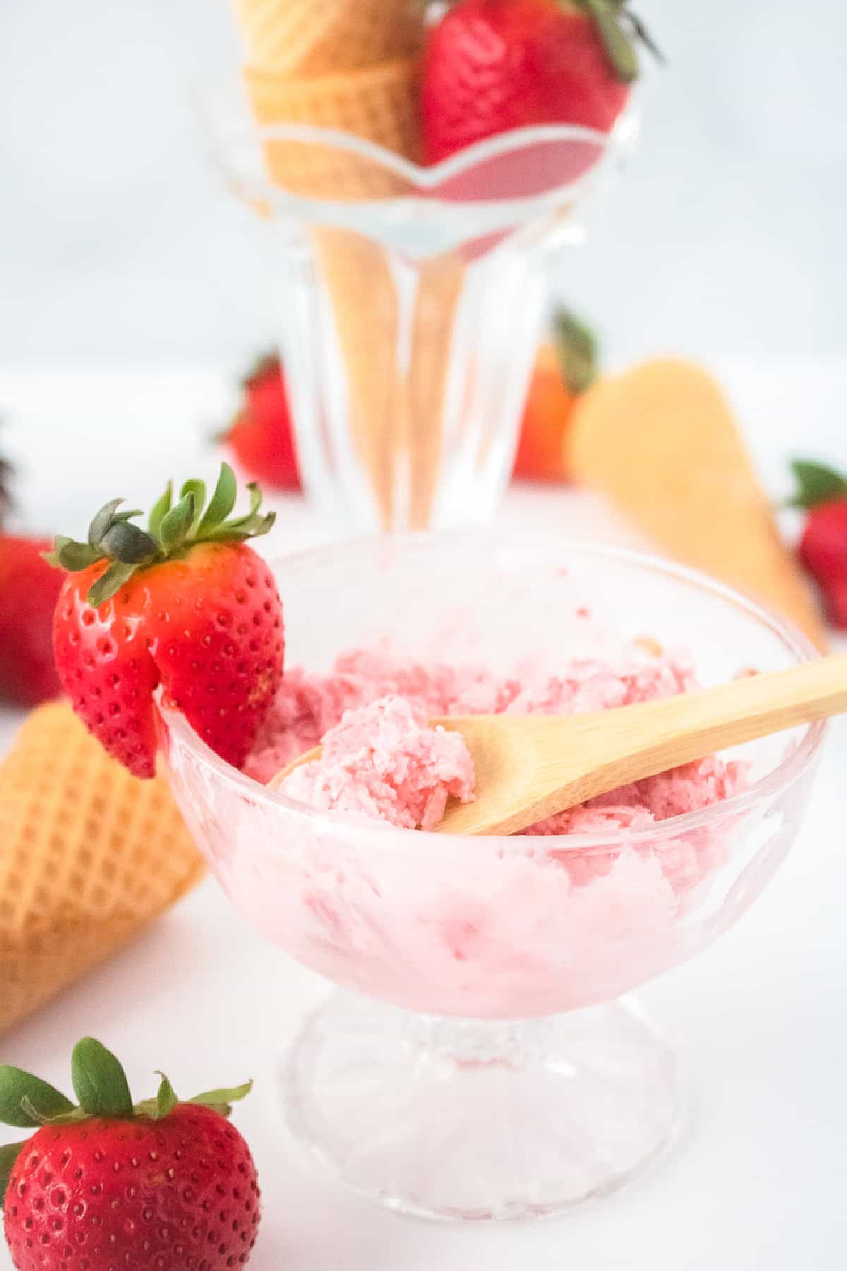 Vegan strawberry ice cream served in a glass bowl with wooden spoon and fresh strawberries in the background.