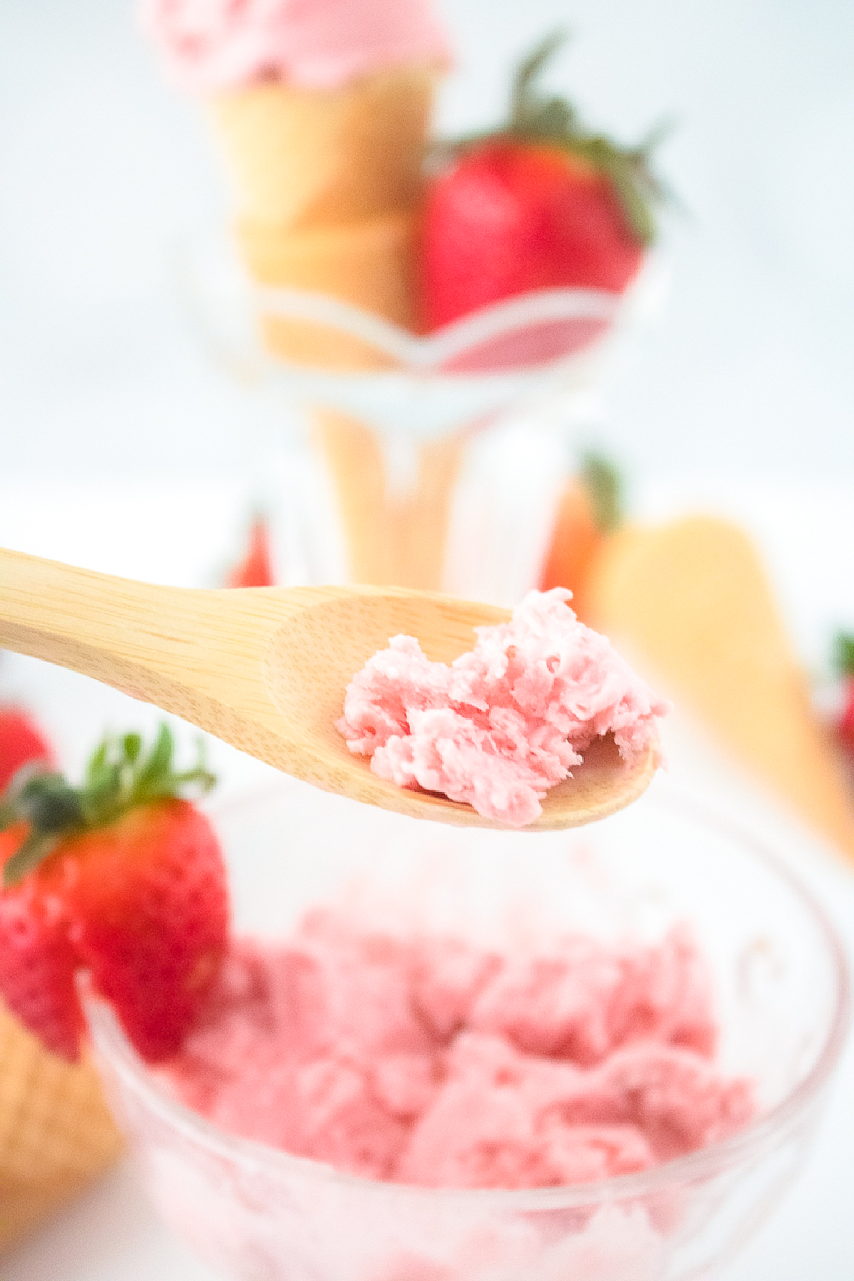 Wooden spoon holding a bite of vegan strawberry ice cream above a bowl of ice cream with waffle cones and strawberries around.