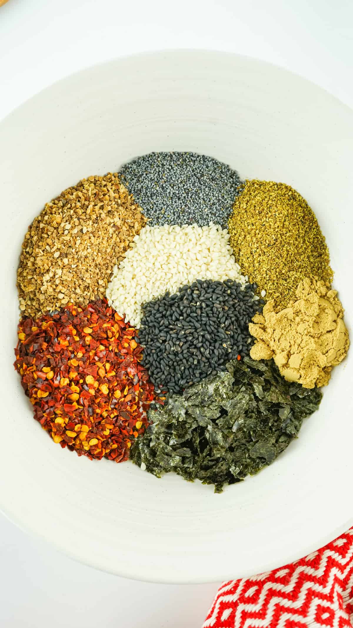 A white bowl filled with neatly arranged spices for Japanese 7 spice blend, including sesame seeds, chili flakes, seaweed flakes, ginger powder, and more, ready to be mixed.