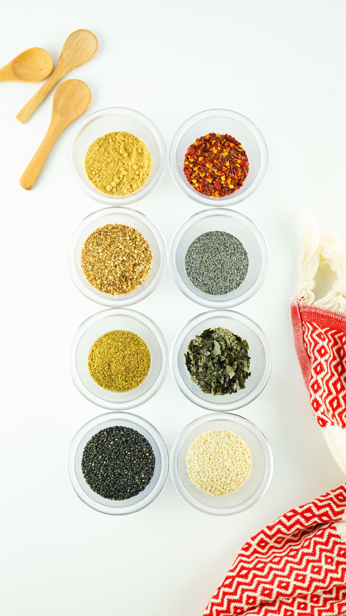 Eight small bowls filled with spices for Japanese 7 spice seasoning arranged in a neat grid on a white background with wooden spoons and a red patterned cloth.