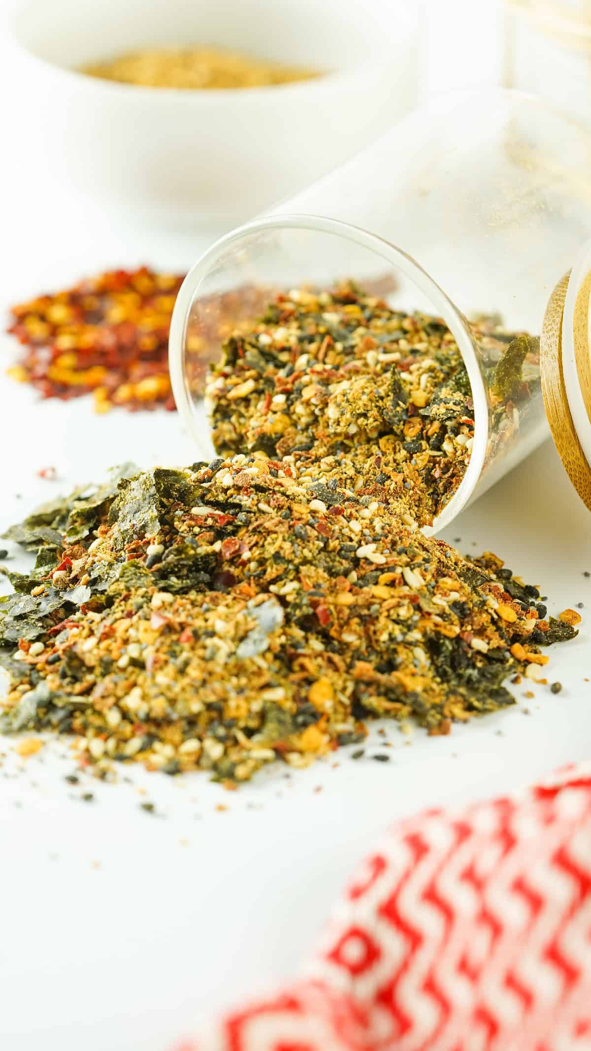 Homemade Japanese 7 spice blend spilling from a tipped glass jar onto a white surface with scattered chili flakes.