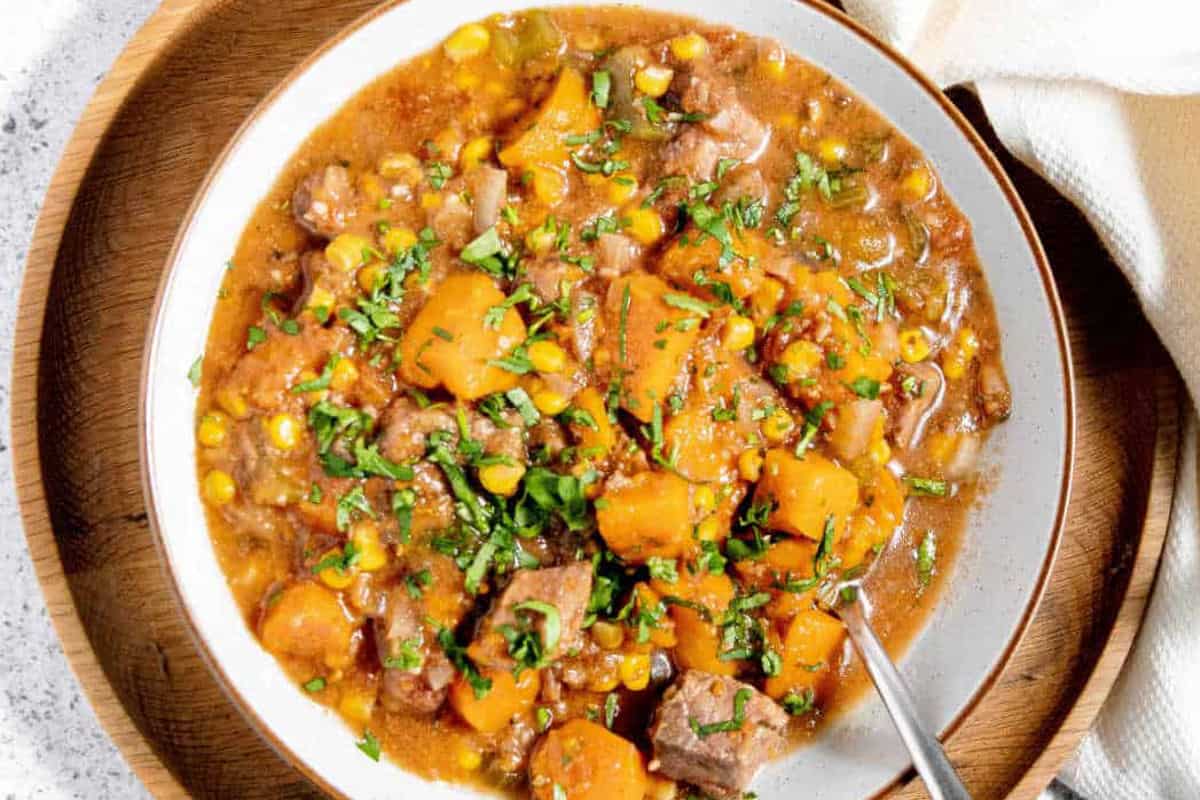 A bowl of beef stew with chunks of beef, carrots, potatoes, and corn, garnished with herbs. A spoon rests in the bowl, and a cloth napkin is nearby on a wooden plate.