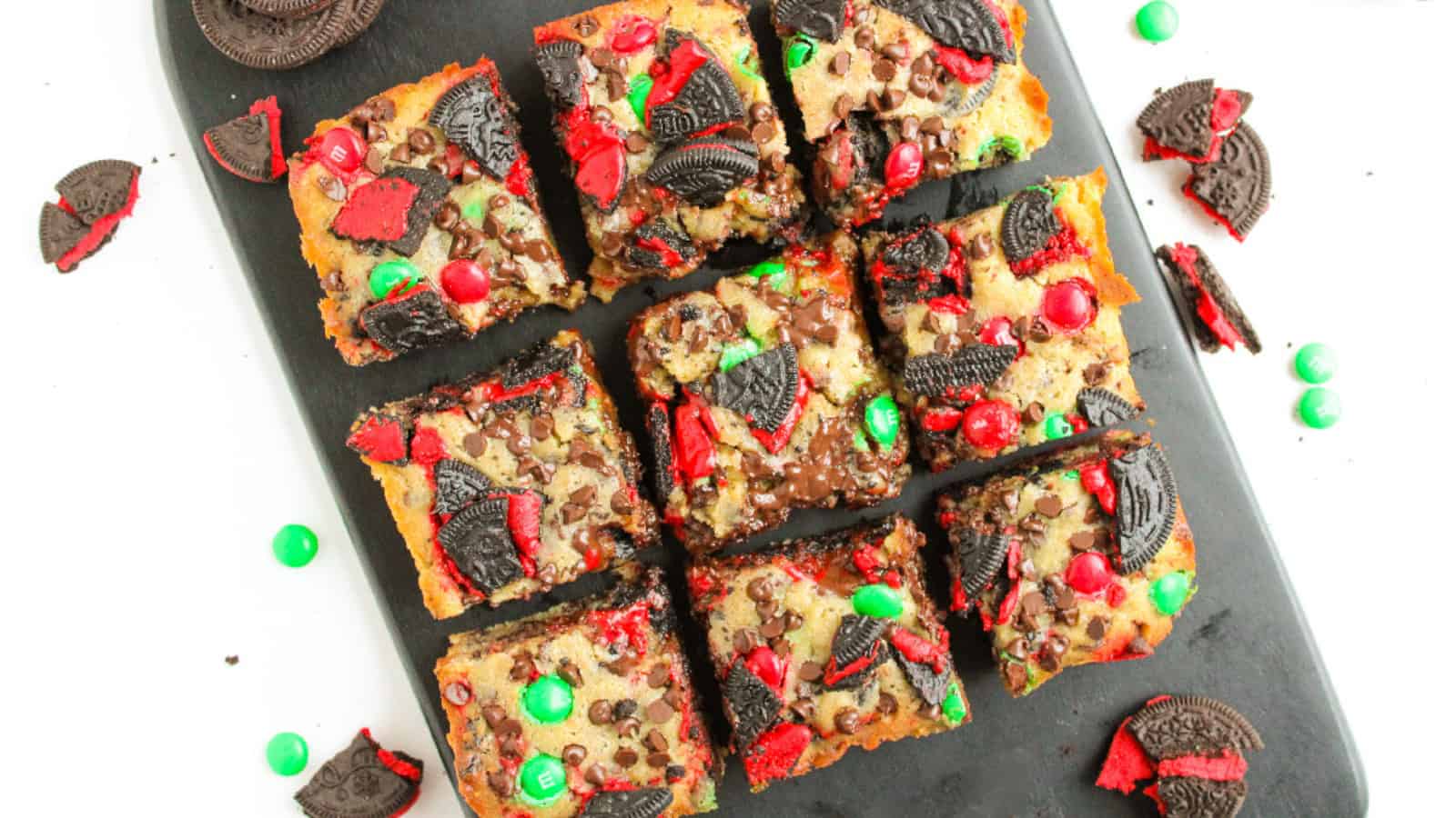 Top-down view of Christmas Oreo cookie bars cut into squares, topped with red and green M&Ms, crushed red-filled Oreos, and mini chocolate chips on a black serving board.