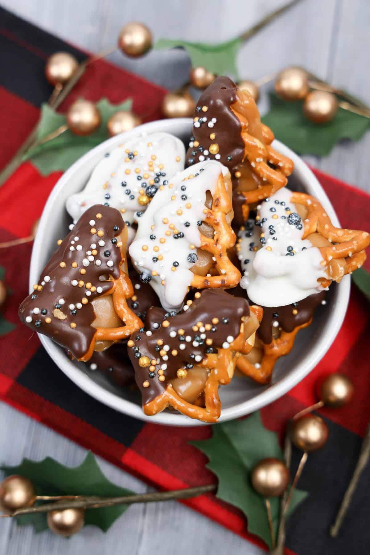 Festive caramel pretzel candies in a bowl, dipped in chocolate and sprinkled with colorful holiday pearls.