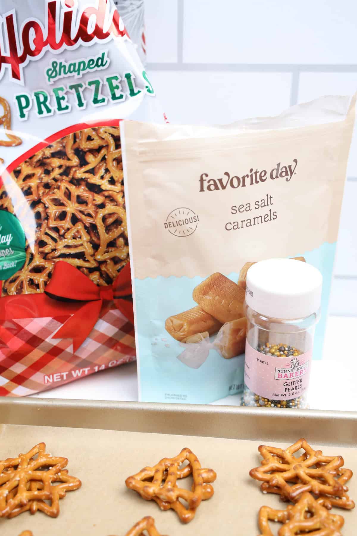 Ingredients for caramel pretzel bites, including holiday-shaped pretzels, sea salt caramels, and festive sprinkle pearls on a baking sheet.