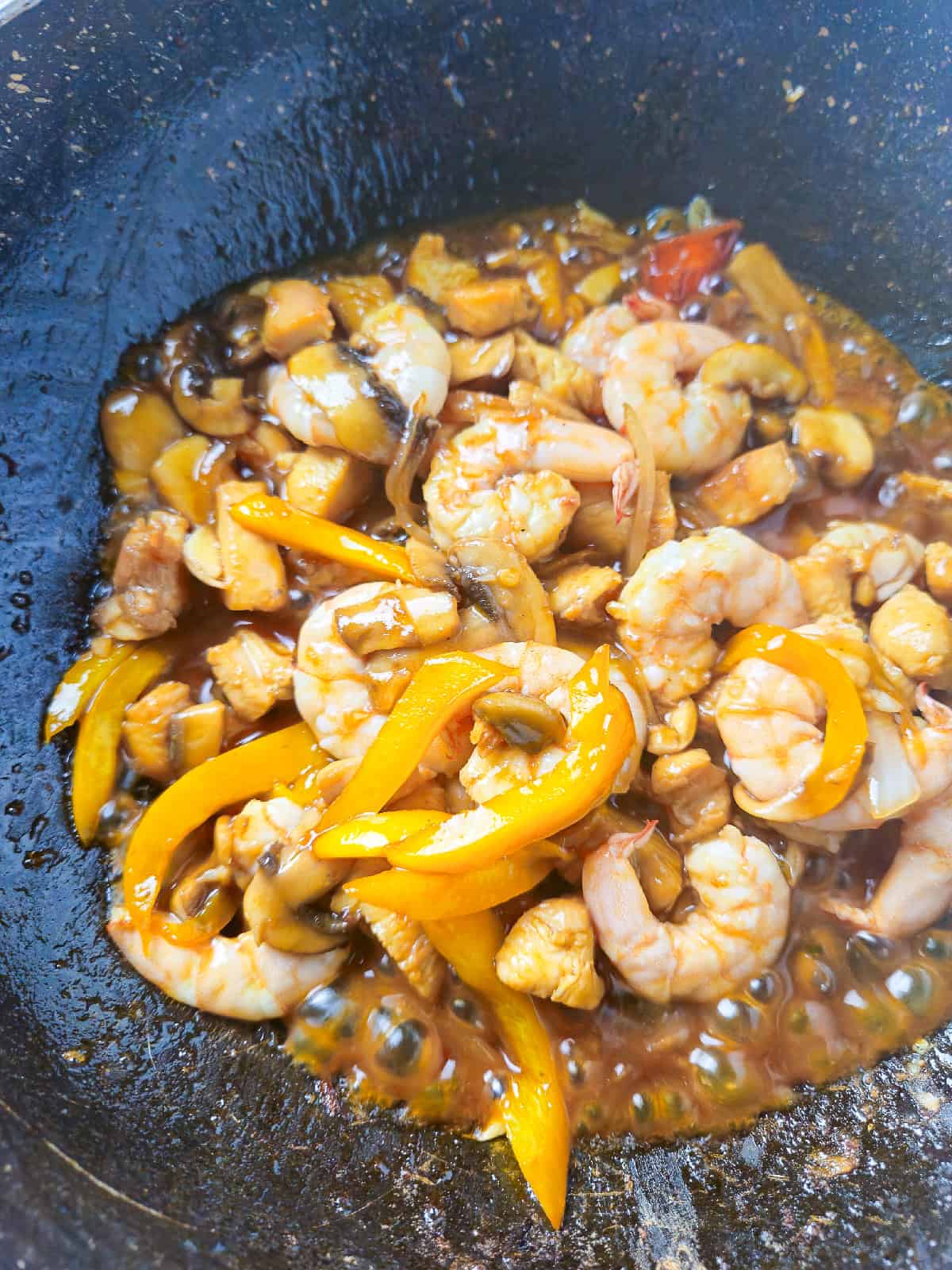 Shrimp, chicken, mushrooms, and bell peppers simmering in sauce inside a hot wok.
