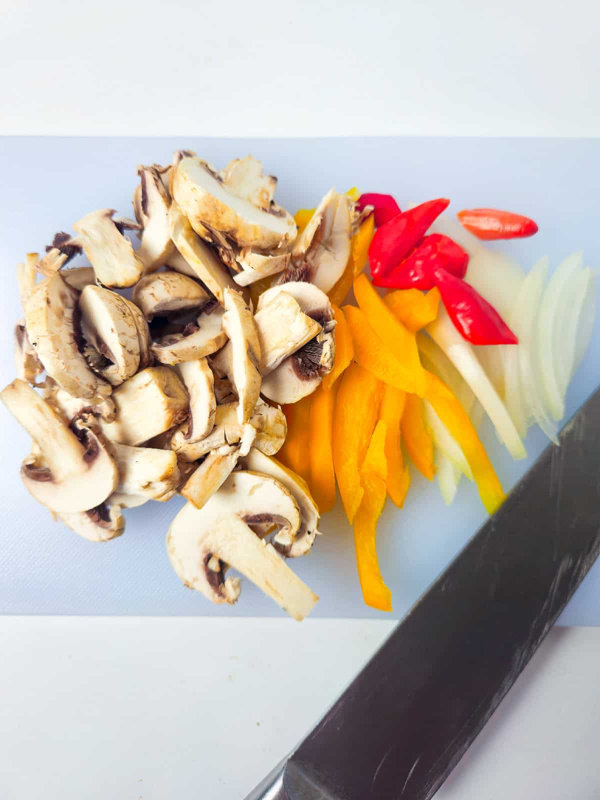 Fresh sliced mushrooms, bell peppers, chili peppers, and onions on a cutting board with a knife.