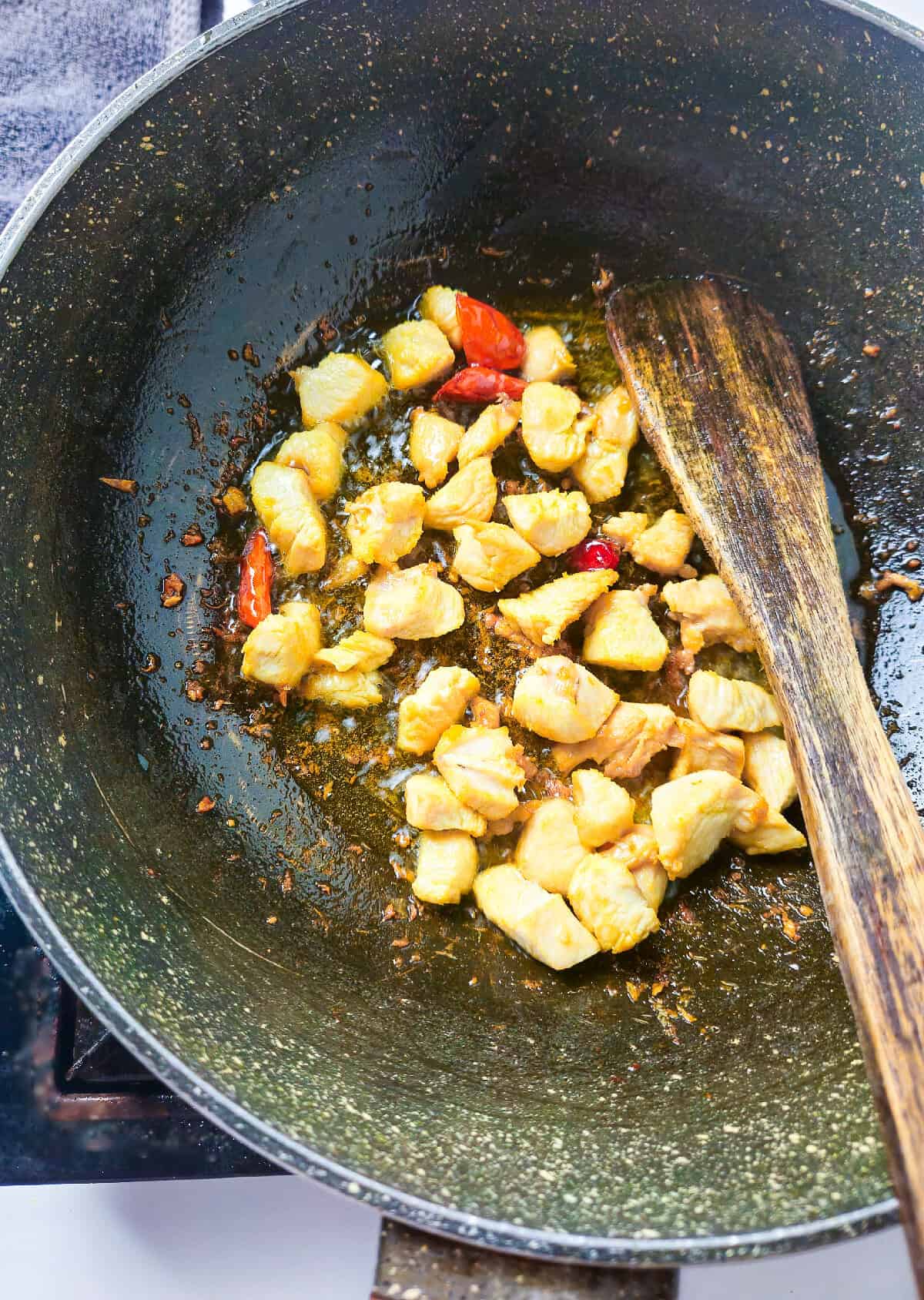 Browned chicken pieces sautéing with chili peppers in a wok using a wooden spatula.