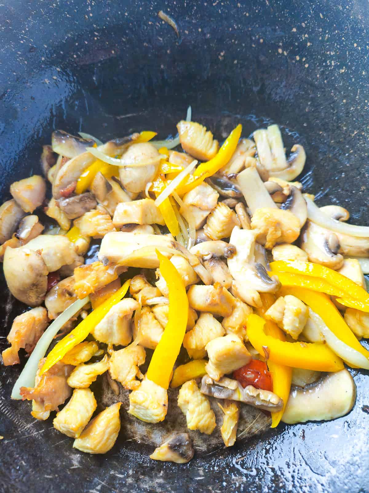 Chicken pieces cooking with mushrooms, onions, and yellow pepper inside a wok.
