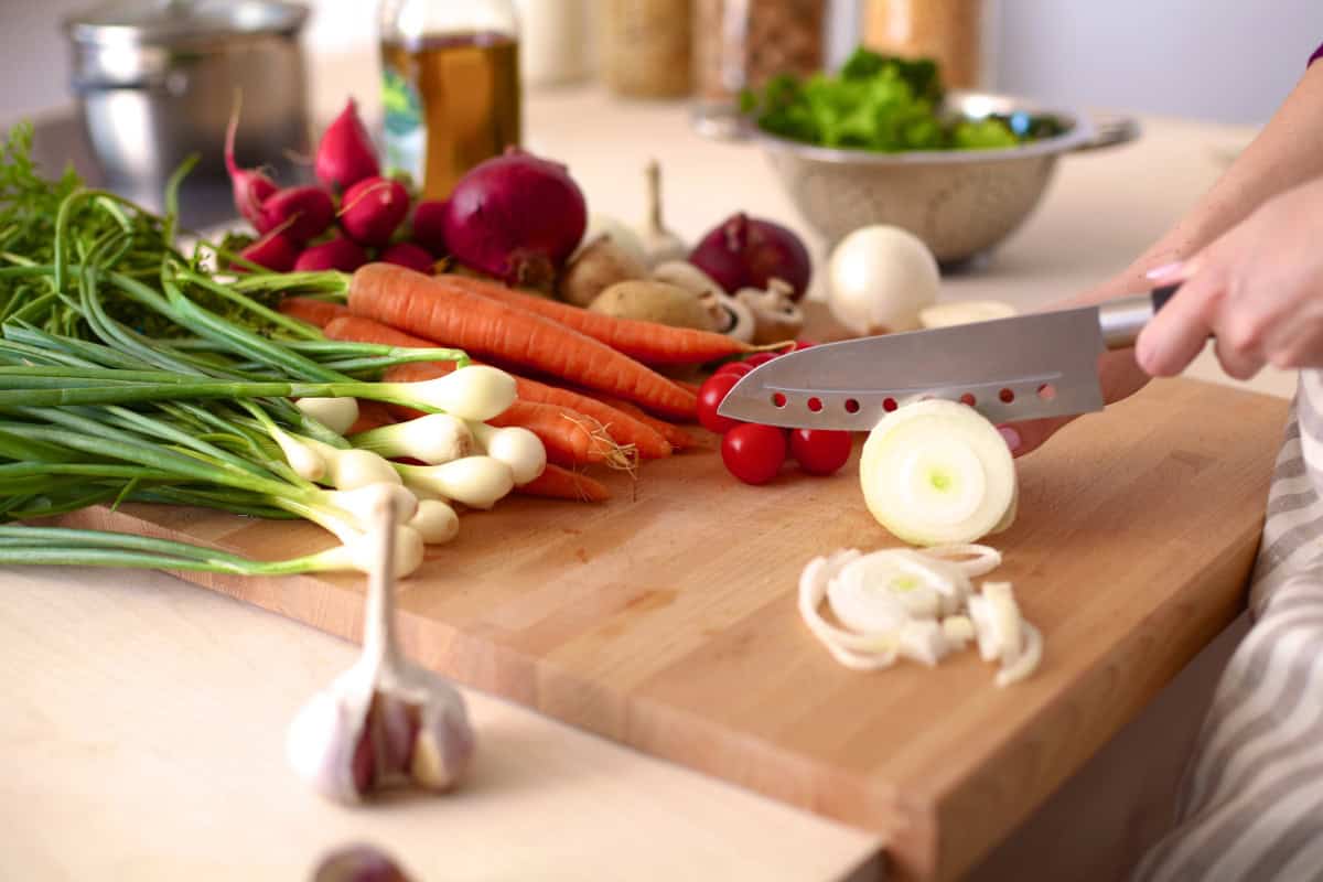 Hands slicing an onion on a wooden cutting board surrounded by fresh vegetables like carrots, green onions, radishes, and cherry tomatoes.