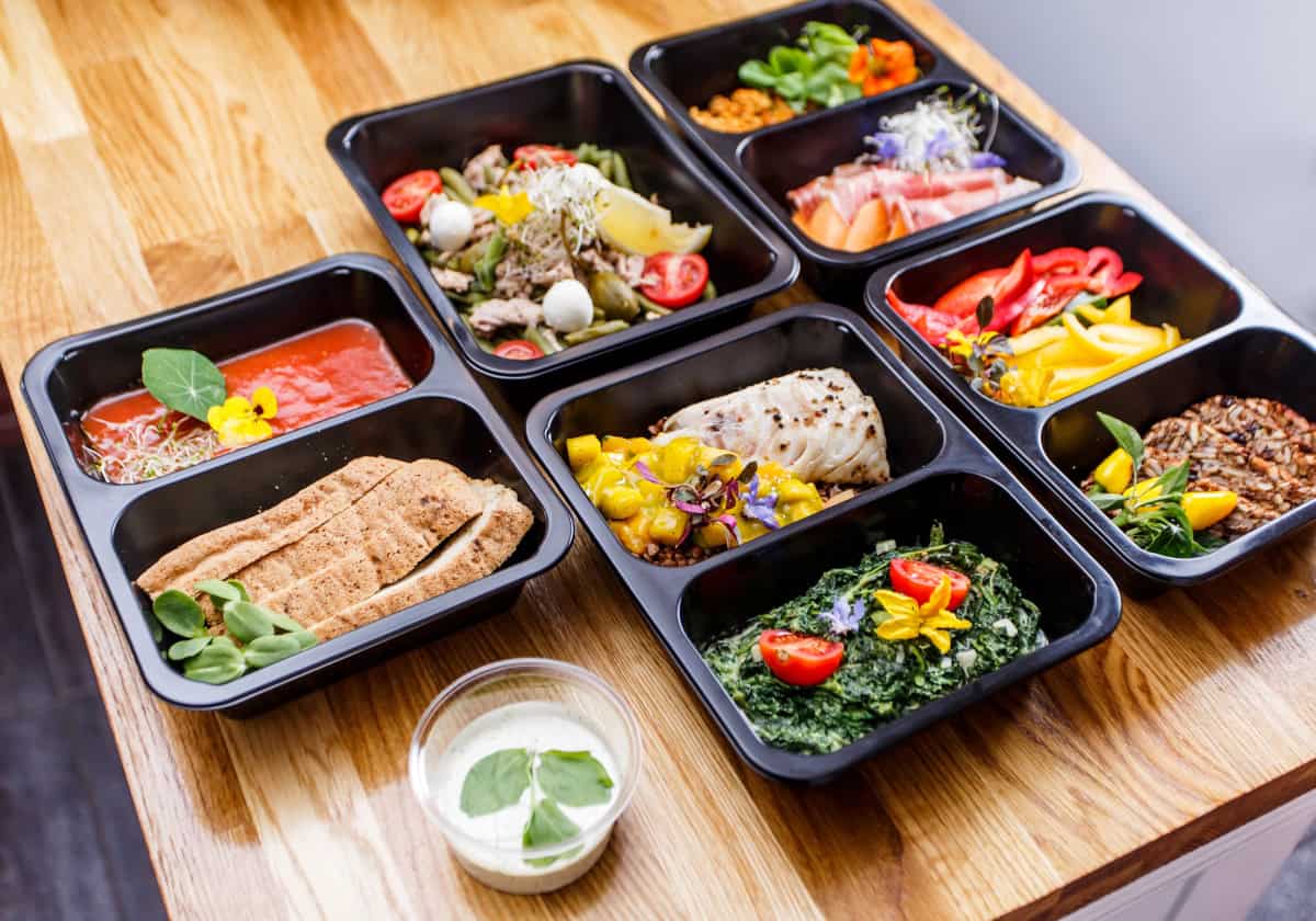 Assorted healthy meal prep dishes arranged in black containers, featuring fish, vegetables, grains, and sauces on a wooden table.