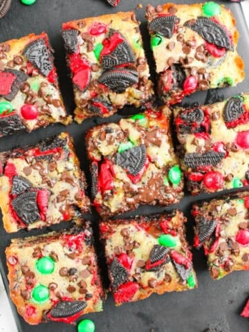 Top-down view of festive Christmas Oreo cookie bars cut into nine squares, filled with red and green M&M’s, crushed Oreo pieces, and melted chocolate on a black serving board.