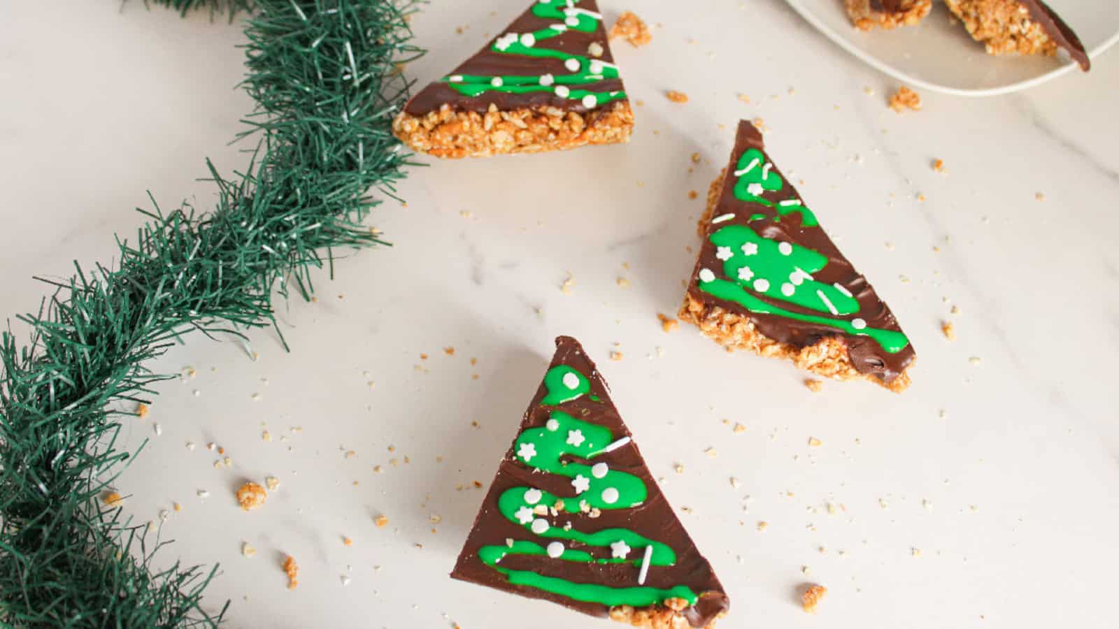 Three Christmas tree&ndash;shaped peanut butter bars arranged on a marble surface with green garland decoration, topped with chocolate and festive green icing with white sprinkles.