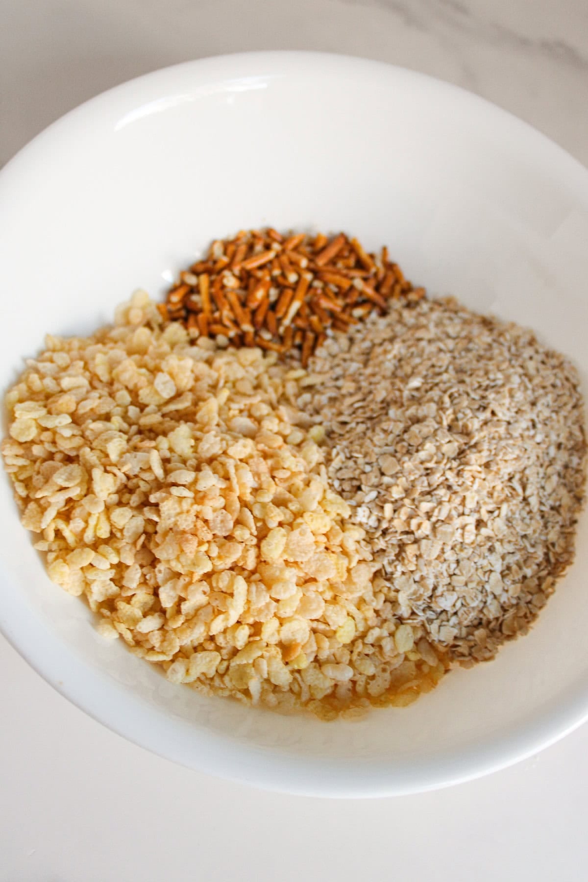 A white mixing bowl filled with crispy rice cereal, crushed pretzels, and oats before mixing.