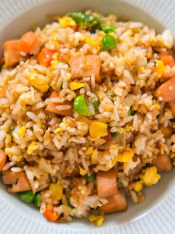 Colorful bowl of spam fried rice mixed with peas, carrots, corn, and scrambled eggs, served in a light gray plate.
