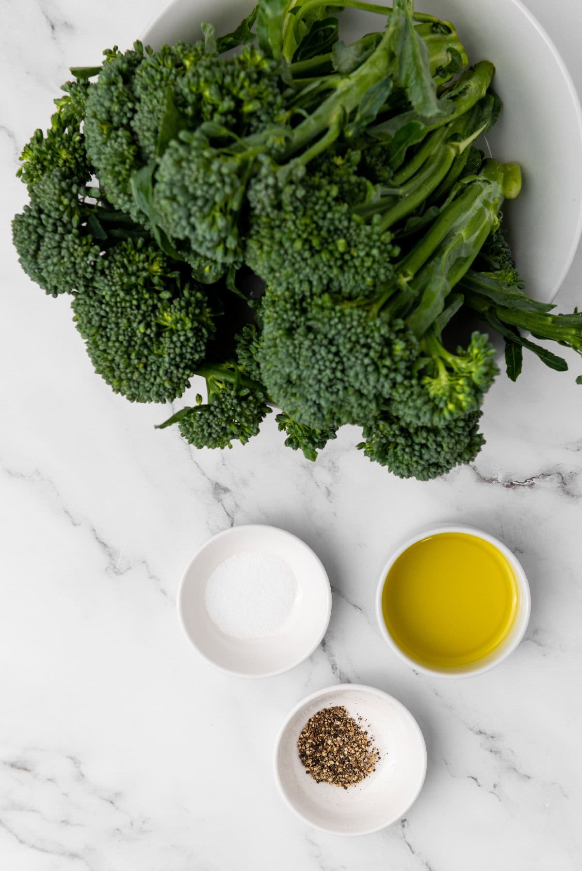 Fresh broccolini on a white plate with bowls of olive oil, salt, and black pepper on a marble surface.