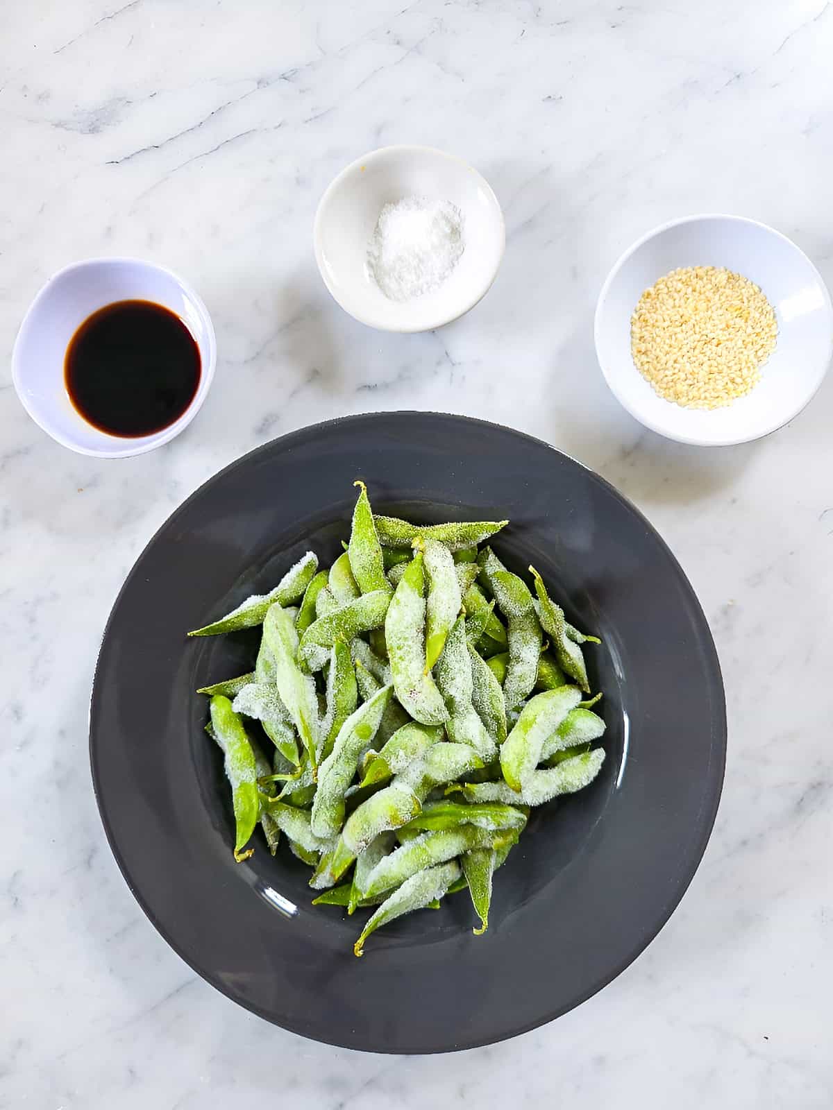 Frozen edamame in a bowl with soy sauce, salt, and sesame seeds on a marble surface.